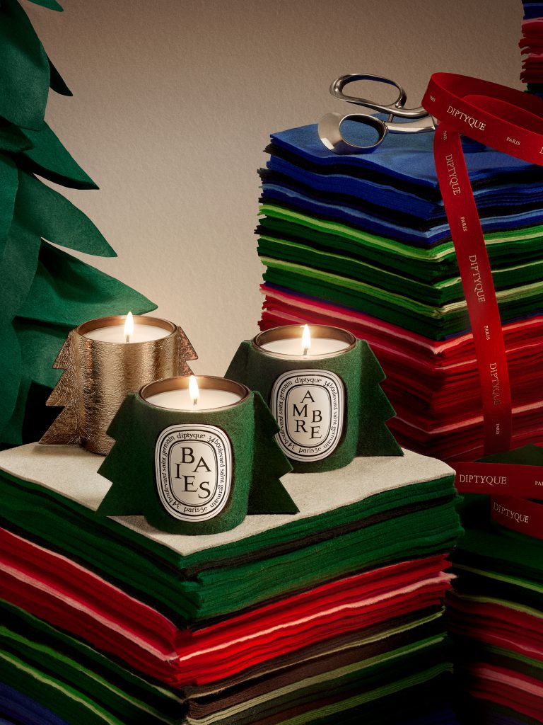 Hai ngọn nến Diptyque đặt tr&ecirc;n chồng giấy m&agrave;u đỏ xanh với k&eacute;o v&agrave; trang tr&iacute; Gi&aacute;ng sinh