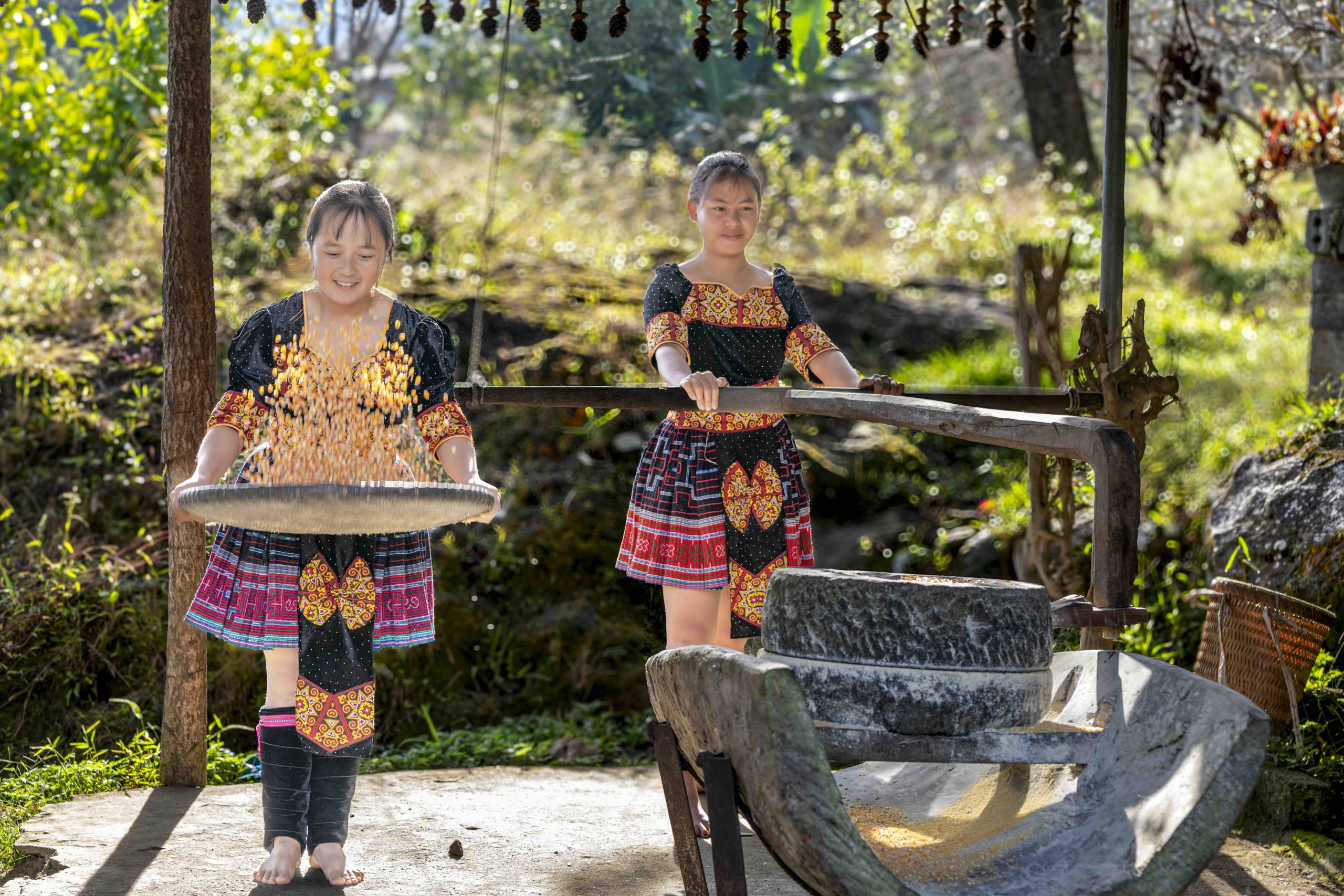 Two young women in colorful clothing process grain with a stone mill outdoors